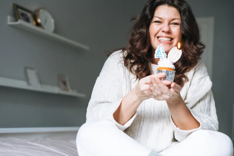 young adult happy beautiful woman forty years plus size body positive in white cozy knitted sweater with festive cupcake with candle in hands on bed at the home party, birthday girl