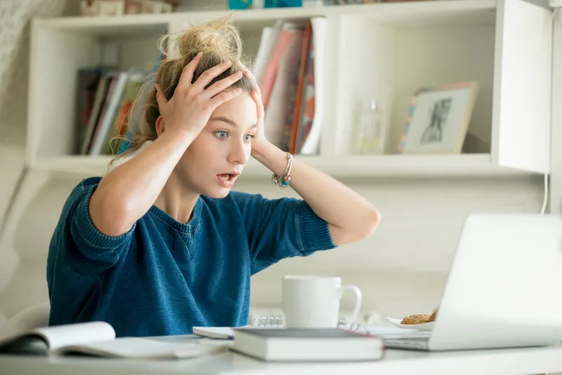 portrait of an attractive woman at the table with cup and laptop, book, notebook on it, grabbing her head bookshelf at the background, concept photo
