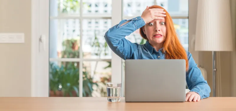 redhead woman using computer laptop at home stressed with hand on head, shocked with shame and surprise face, angry and frustrated fear and upset for mistake