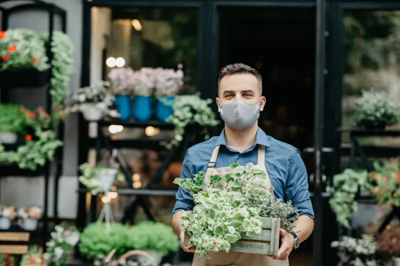 small business and start of working day man in protective mask takes out box of plants outside in front of flower shop