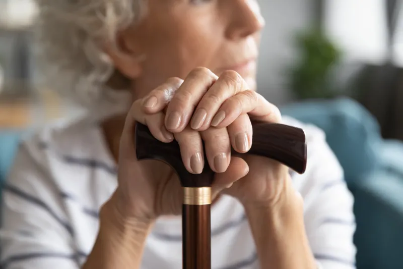 close up focus on wrinkled female hands lying on walking cane disabled middle aged elder woman holding wooden stick, resting sitting on sofa indoors, disability retirement people lifestyle concept