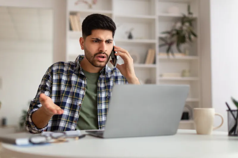 confused discontented arab man talking on phone using laptop having problem with computer or internet connection at home displeased male customer calling to hotline service and complaining