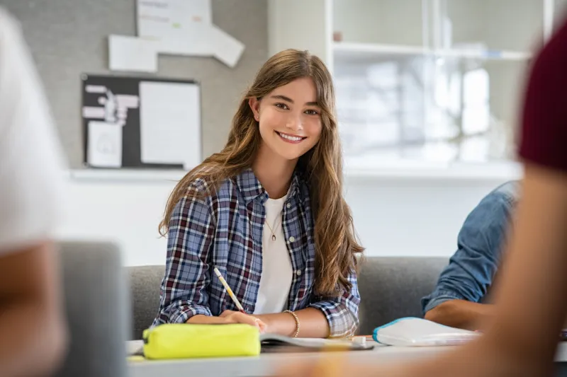 beautiful university student smiling while studying for exams in classroom pretty woman sitting in classroom full of students during class portrait of happy young woman writing notes and looking at camera