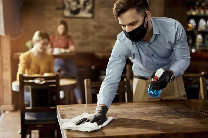 young waiter wearing protective face mask while cleaning tables while working in a cafe
