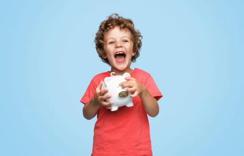 cute little boy with piggy bank laughing and looking at camera while standing on blue background