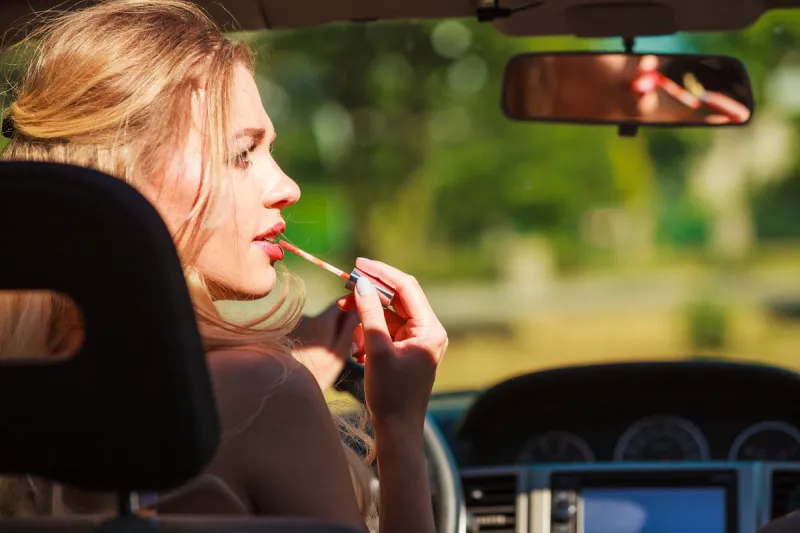 concept of danger auto drive young woman driver painting her lips doing applying make up while driving the car