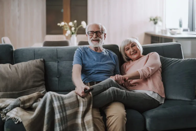 love and tendance in any age front view portrait of happy smiling gray haired man holding legs covering in plaid of her beloved wife while enjoying time together on sofa