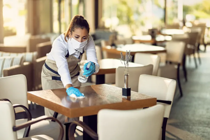 young waitress disinfecting tables while wearing protective face mask ad gloves due to coronavirus epidemic