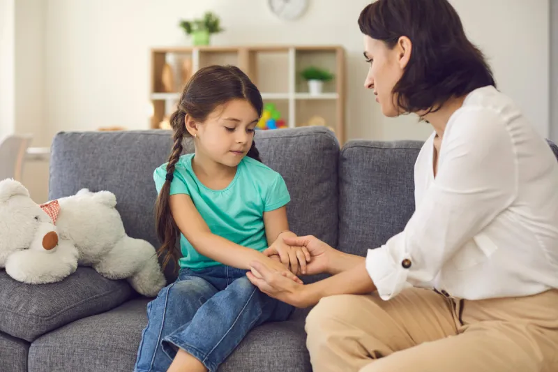 young mother sitting on sofa with daughter, holding her hands and talking to her seriously at home with room interior at background solving problems in children education concept
