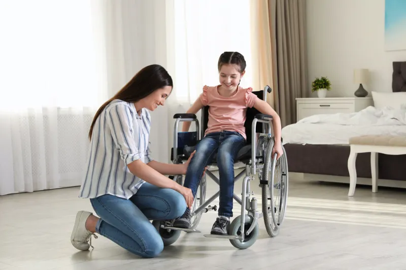 young woman helping her disabled daughter get in wheelchair at home