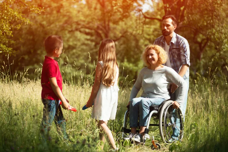 cheerful woman on a wheelchair is laughing with her husband and kids while playing games outdoor happy family concept