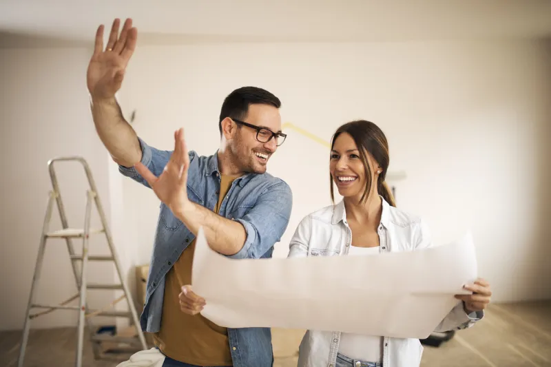 young couple standing in their apartment while woman holding blueprint and man showing to her new ideas