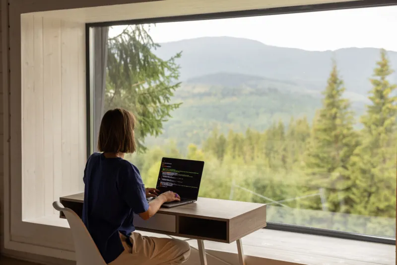woman works on laptop while sitting by the table in front of panoramic window with great view on mountains remote work and escaping to nature concept