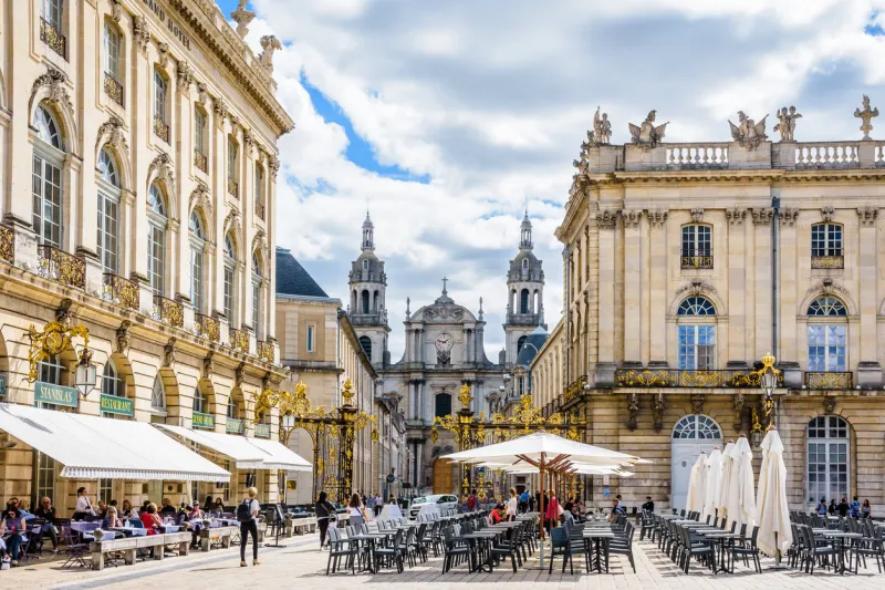 nancy, france - september 12, 2019  sunshine on the terrace of the sidewalk cafe of the grand hotel on the place stanislas closed by a gilded wrought iron gate with nancy cathedral in the background