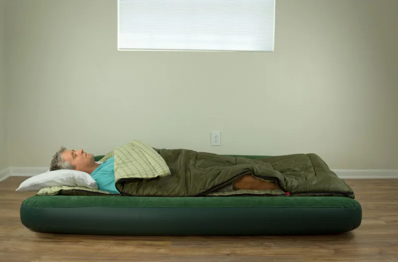man comfortably sleeping on blow up air mattress bed in sleeping bag with clean walls and window in the background