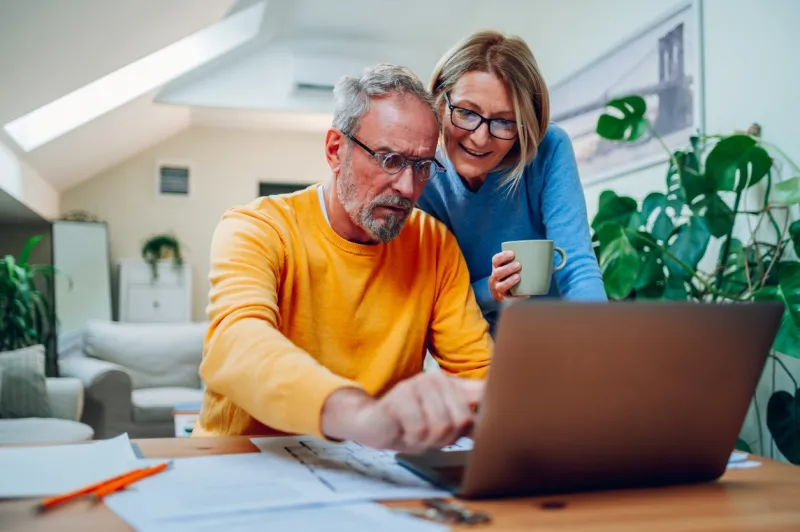 happy senior couple laughing and bonding while using laptop at home smiling elderly husband and wife having fun satisfied with buying insurance, paying bills online man showing something on laptop
