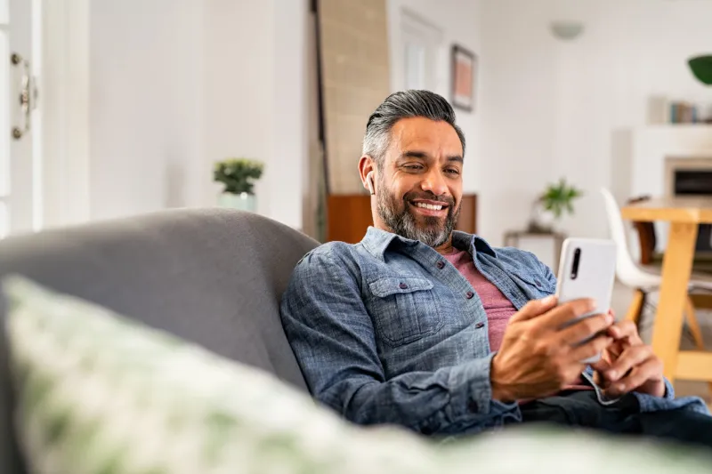 happy mid adult man using smartphone device while sitting on sofa at home smiling indian man lying on couch reading messages on mobile phone while listening to music with wireless earbuds mature guy relaxing at home