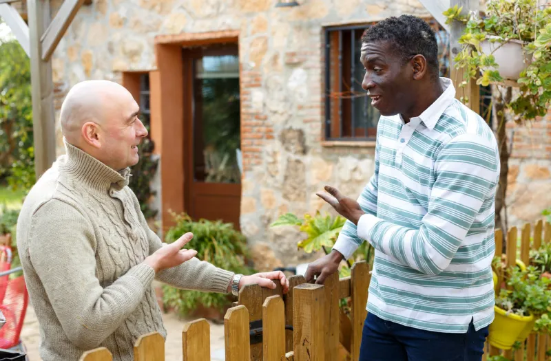 two male farmers friendly talking outside next to wooden fence on background with brick house