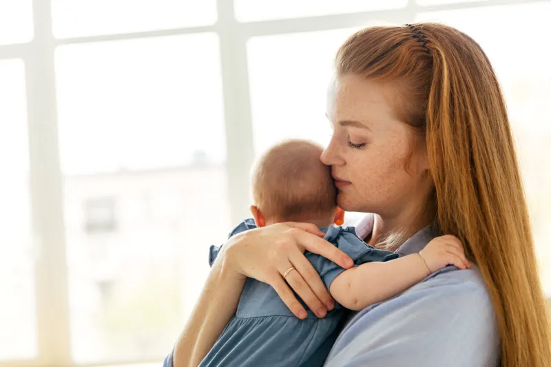 young mother with a three-month-old baby by the window in the bedroom, thoughtful and tired