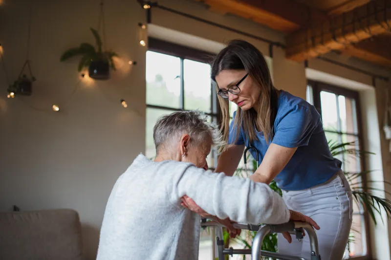 a healthcare worker or caregiver visiting senior woman indoors at home, helping her to walk