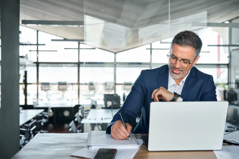 middle aged smiling professional business man global company executive ceo manager or lawyer wearing suit sitting at desk in modern office working on laptop computer and writing notes, copy space