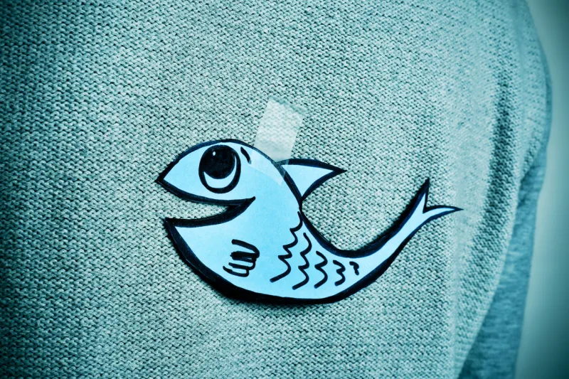 closeup of a young man with a paper fish attached with tape to his back, with a slight vignette added