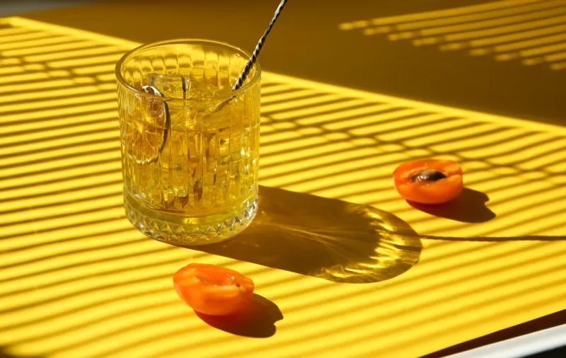 drinks and cocktails minimalism whiskey with ice in a clear glass with a bartender's spoon, lime chips and fresh apricot on a yellow background sunny bright light background image, copy space