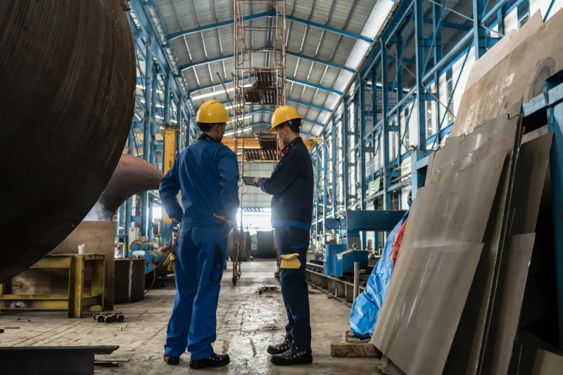 two workers wearing yellow hard hat and blue uniform in the interior of an industrial hall