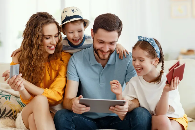 smiling parents with kids gathering on sofa using tablet while picking tour online anticipating summer vacation all together