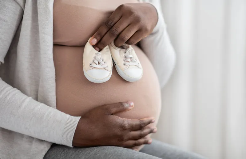 desired pregnancy black pregnant woman embracing belly and holding small baby shoes near her tummy, expectant lady enjoying maternity time, cropped image with selective focus, closeup shot