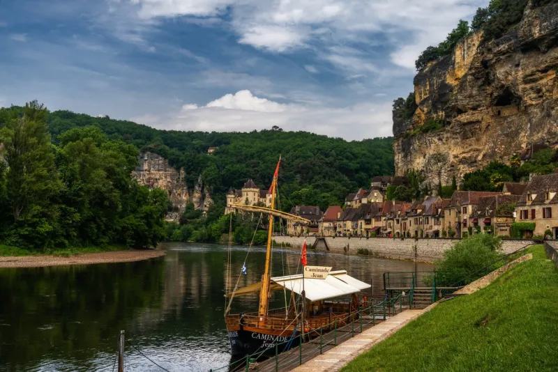 la roque-gageac, france - 12 may, 2022  view of the historic village of la roque-gageac with the dordogne river and cliffside houses and a wooden gabare river boat in the foreground