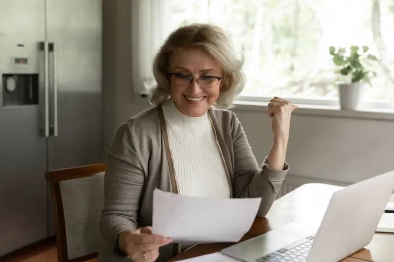 happy adult retiree woman reading bank document, checking account balance, smiling at investment income, savings or insurance balance, getting good news female pensioner doing paper work at home
