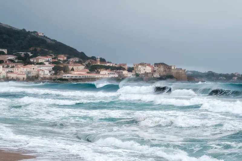 rough seas crash onto the beach at algajola in the balagne region of corsica with village and citadel in the distance