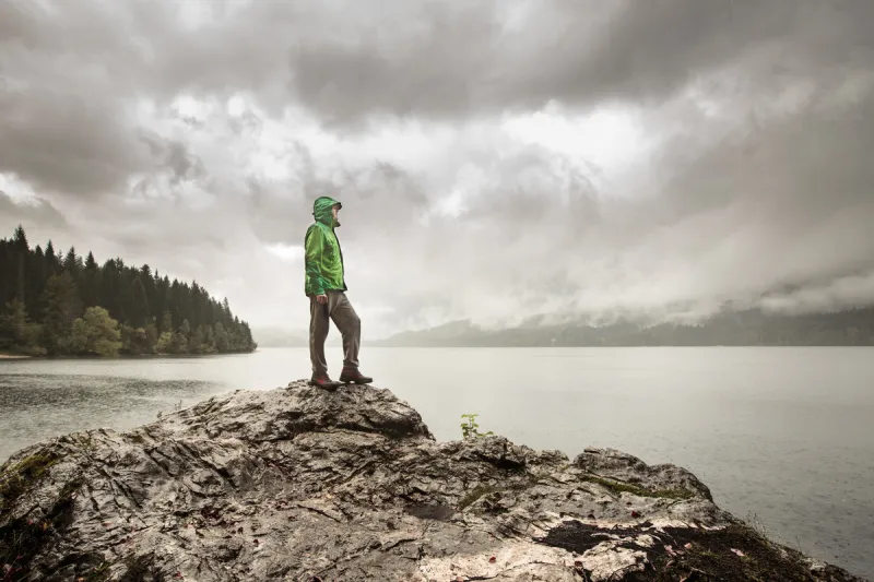 man standing on a rock beside a dramatic mountain lake after a hike in the rainy, gloomy day active lifestyle, outdoor activities, moods and emotions concept