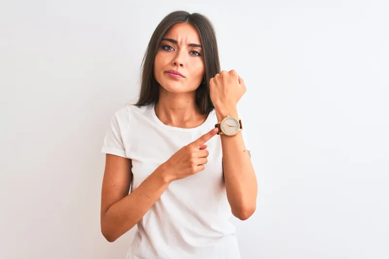 young beautiful woman wearing casual t-shirt standing over isolated white background in hurry pointing to watch time, impatience, looking at the camera with relaxed expression