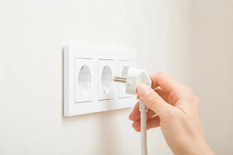 young adult woman hand holding and plugging white electrical plug in wall outlet socket at home closeup side view