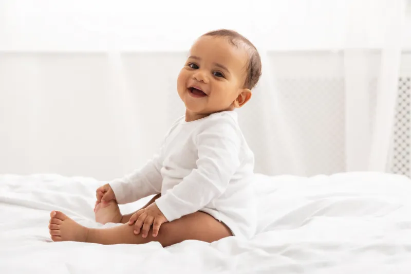 portrait of adorable african american baby toddler sitting on bed and smiling looking at camera posing in bedroom at home happy childhood and child care concept