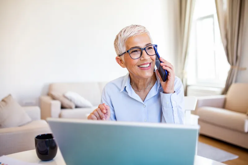 smiling mature beautiful business woman with white hair working on laptop in bright modern home office business woman talking on her mobile phone while working from home