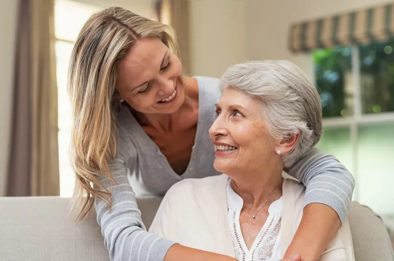 portrait of old mother and mature daughter hugging at home happy senior mother and adult daughter embracing with love on sofa cheerful woman hugging from behind older mom and looking at each other