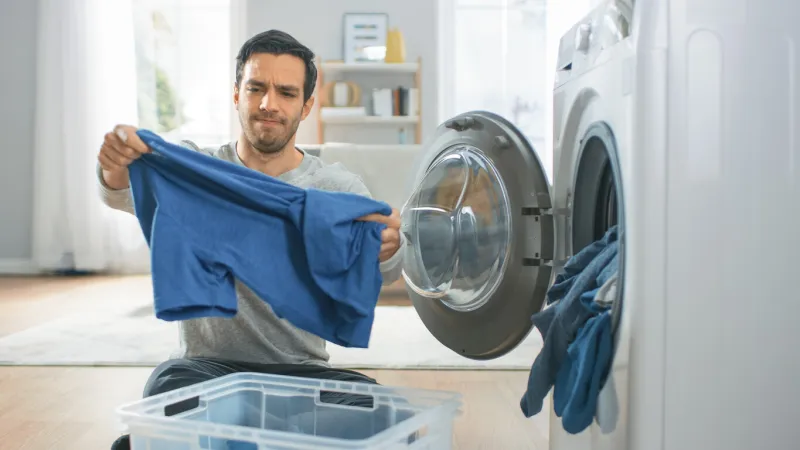 handsome confused young man in grey jeans and coat sits in front of a washing machine at home he loads the washer with dirty laundry bright and spacious living room with modern interior
