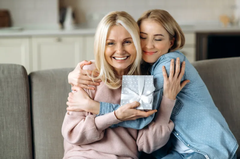 surprise for mother's day or birthday loving young adult daughter giving a present to her beloved middle-aged caucasian mom sitting on the sofa in the living room