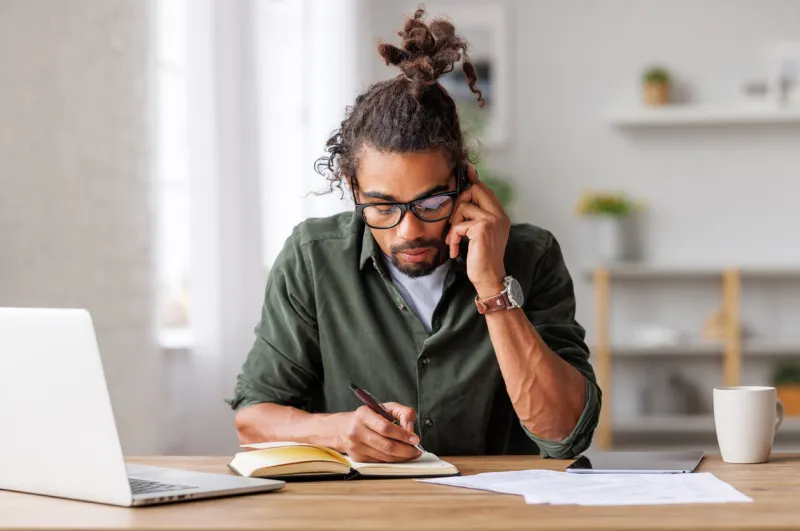 young focused busy man entrepreneur making call via smartphone and writing down notes while working remotely from home on laptop, ethnic guy freelancer talking on mobile phone during remote work