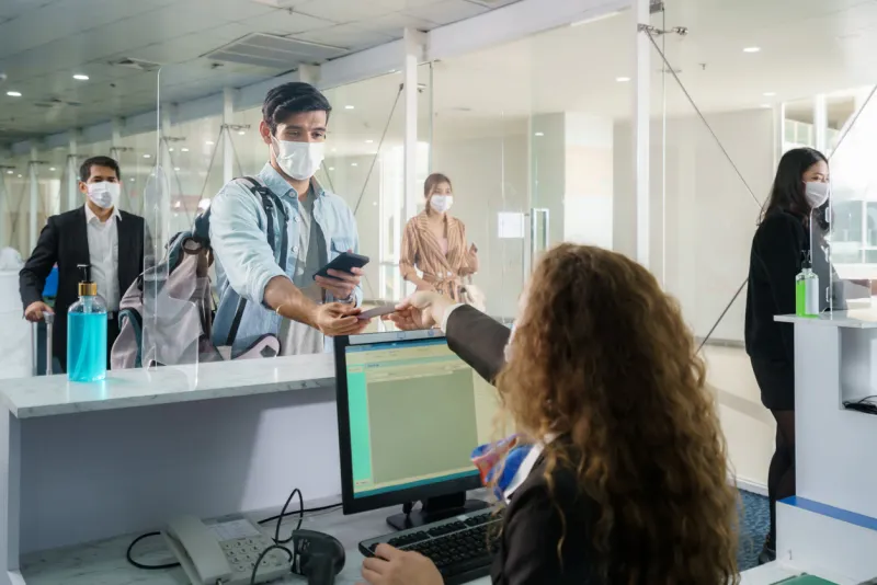 a male airline passengerwith mask is handing over his passport at the airline counter check in through an acrylic barrier for disease prevention coronavirus or covid-19 at airport for new normal travel