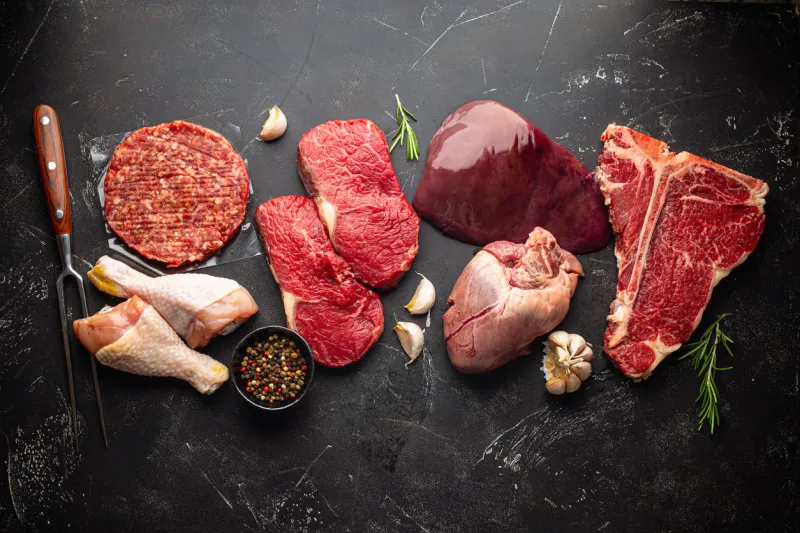 selection of assorted raw meat food with seasonings for zero carb carnivore diet  uncooked beef steak, ground meat patty, heart, liver and chicken legs on black stone background from above