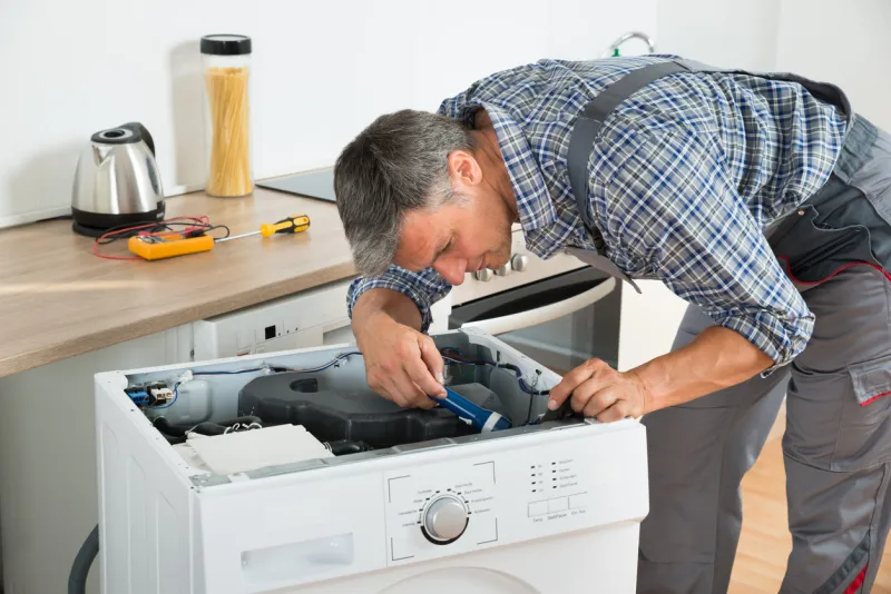 full length of handyman checking washing machine with flashlight in kitchen