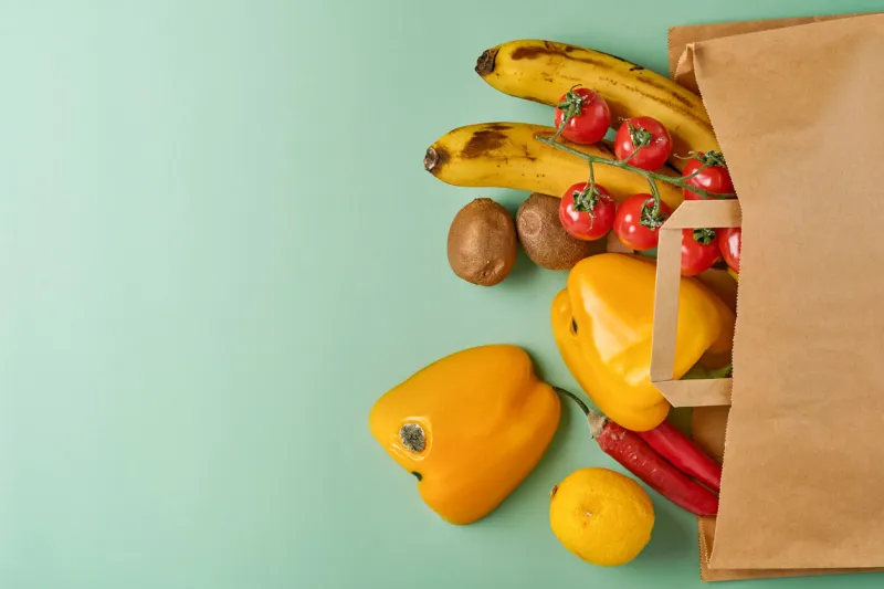 assortment of fresh pepper, chili, bananas, cherry tomato, lemon and kiwi in craft paper bag over green background trendy ugly organic vegetables and fruits top view cooking ugly food concept