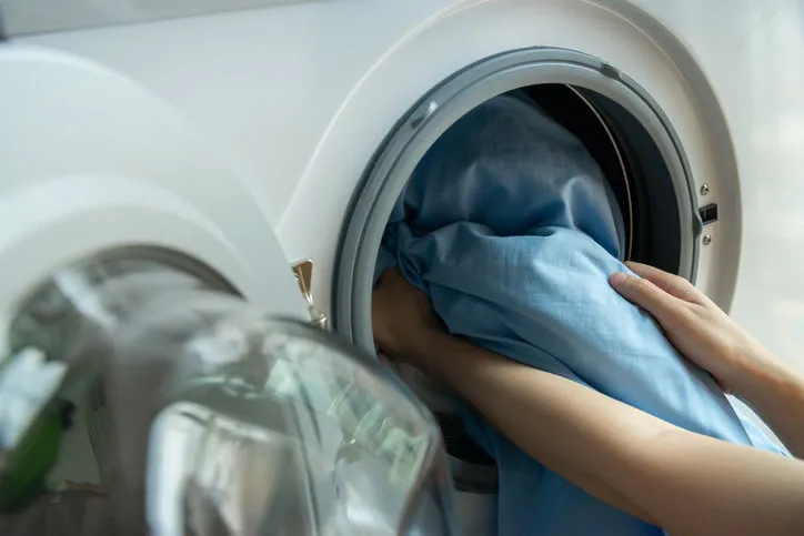 woman’s hand loading dirty blue bed sheets in a white washing machine