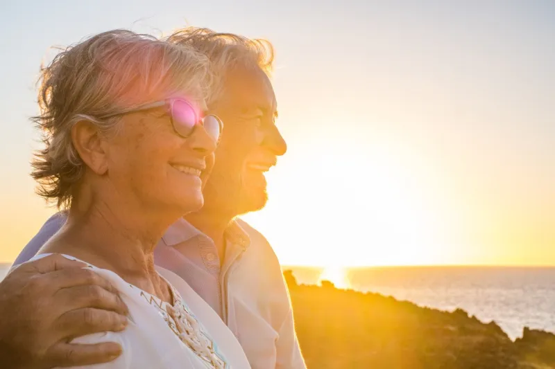 couple of two cute seniors together enjoying summer and having fun at the beach looking at the sea or ocean with sunset - mature people having a good lifestyle
