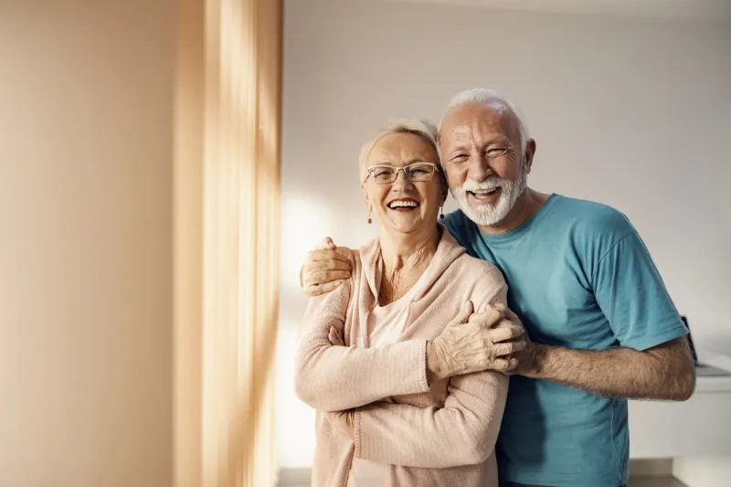 senior couple hugging in a nursing home a happy senior couple standing next to a window in a nursing home, hugging and smiling they have all care they need