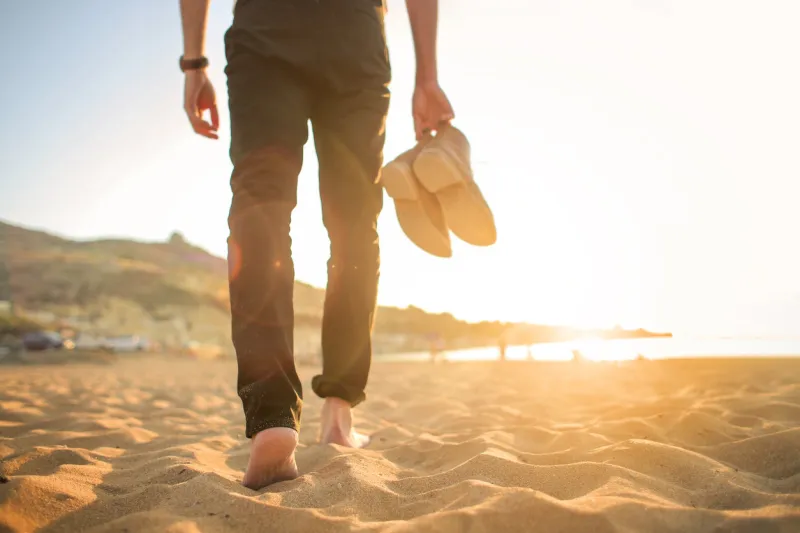 detail of a man walking in the beach, holding a pair of shoes in his hand